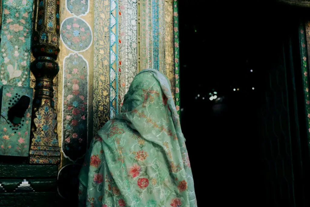 A back view of a woman showing what to wear to a mosque as a non Muslim woman, draped in a light green sheer scarf with red floral embroidery over her head, standing next to an ornately carved and painted turquoise wooden pillars near a dark entrance